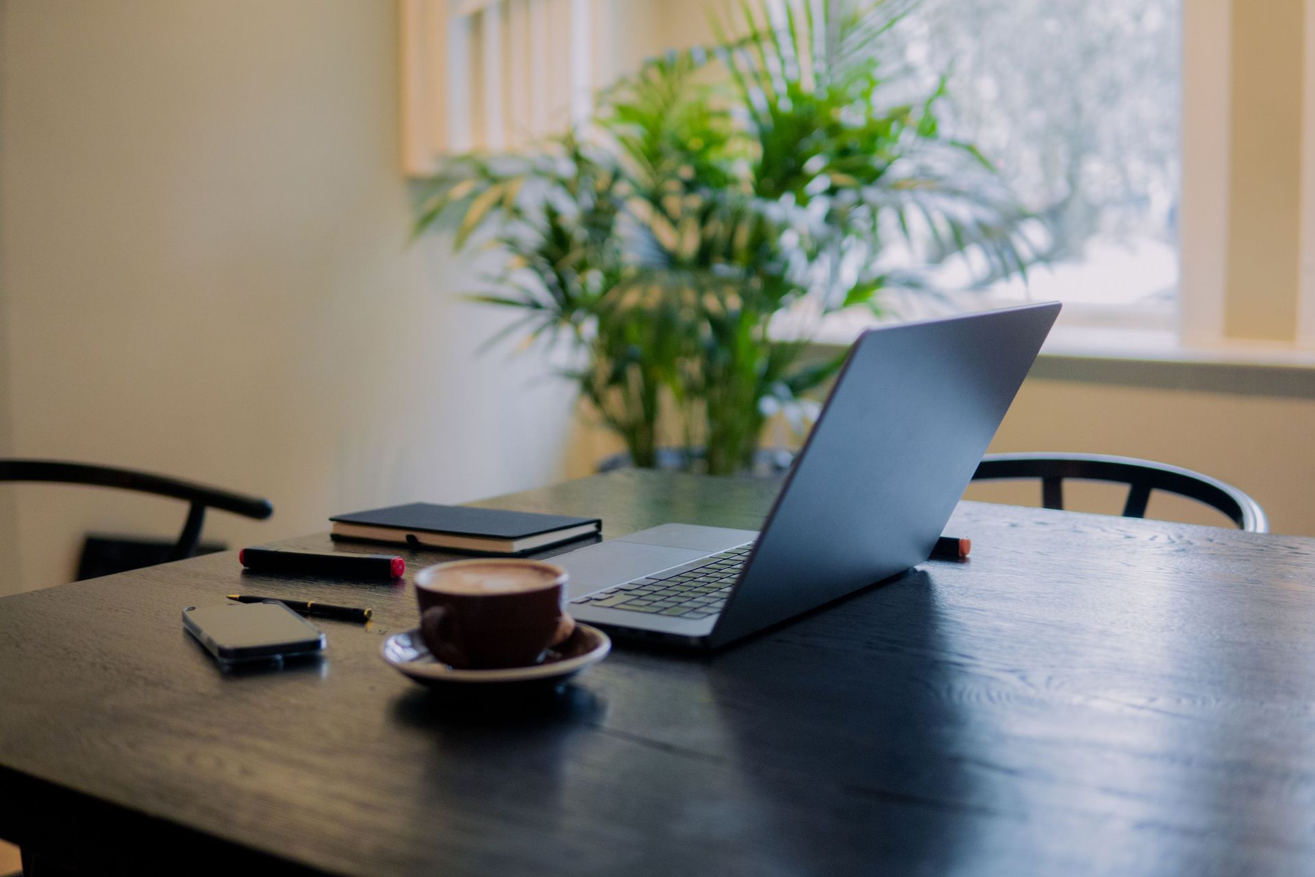 Laptop and coffee cup on a dark table in a bright office with a potted plant nearby