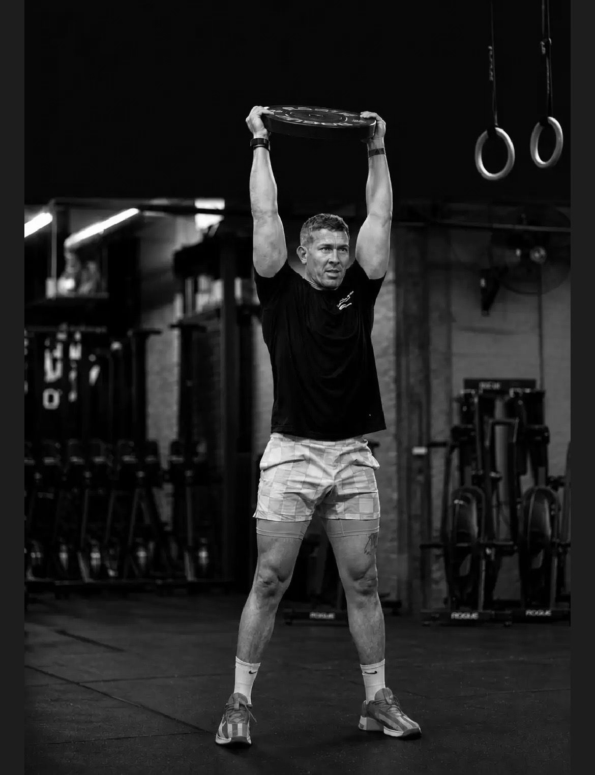 Man in Rittenhouse Fitness, a gym in Philadelphia, PA holding a weight overhead, with rings hanging in the background.