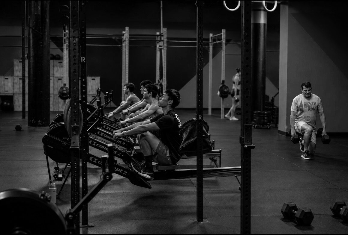 Black-and-white gym scene with people rowing on machines and a man walking nearby