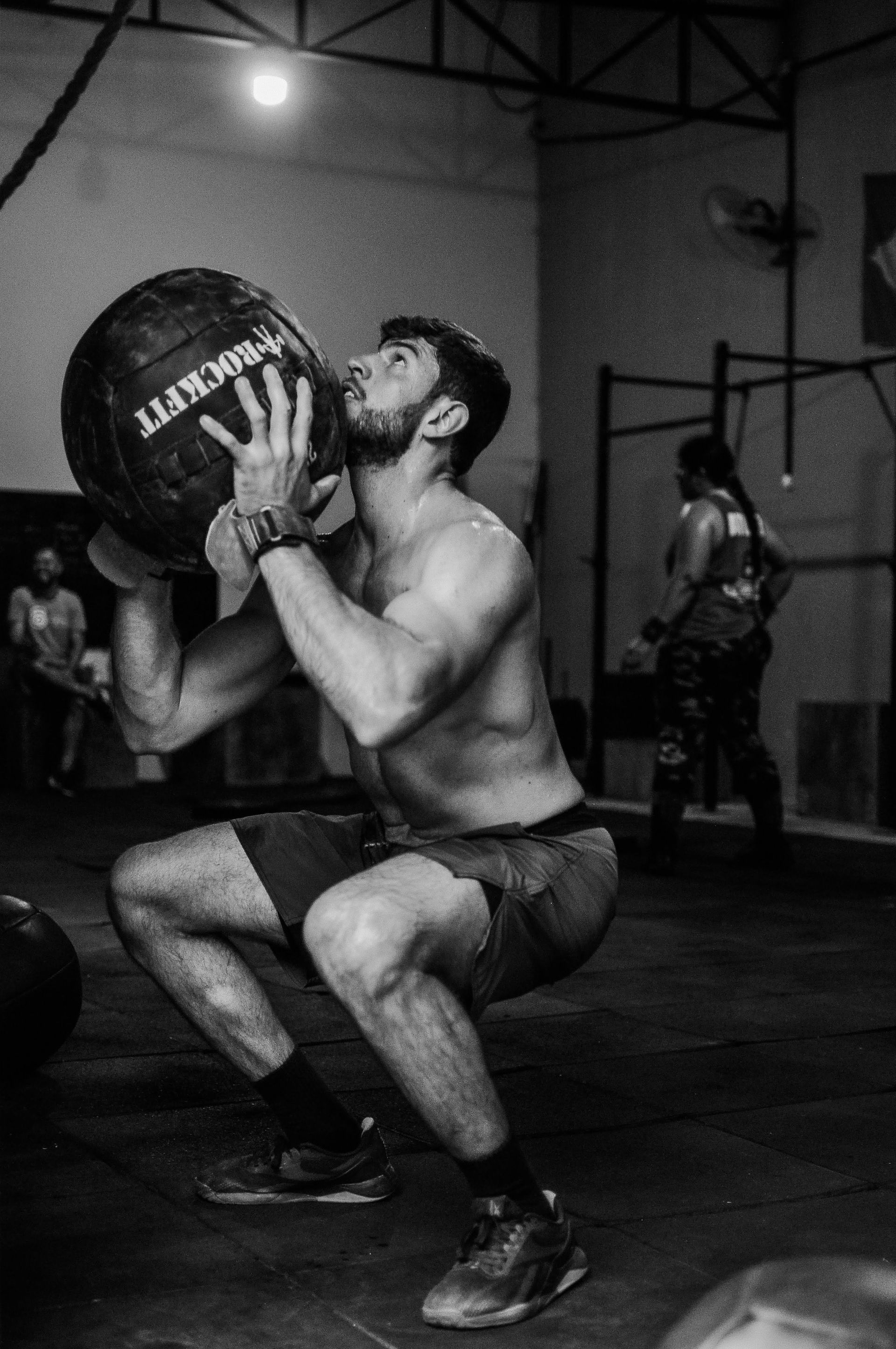 Man squatting and lifting a medicine ball in a gym, with another person in the background.