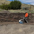 Worker in orange jacket operating a plate compactor beside a muddy roadside embankment.