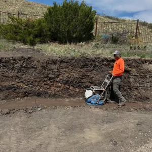 Worker in orange jacket operating a plate compactor beside a muddy roadside embankment.
