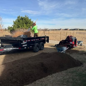 Person in a green shirt loading dark soil into a black trailer beside a wheelbarrow in a rural yard.