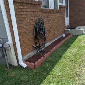 Coiled garden hose mounted on a brick house wall beside a grassy yard and white downspout.