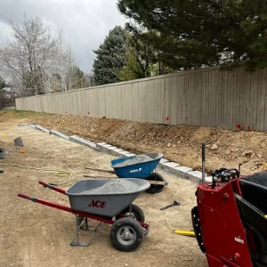 Construction site with wheelbarrows and tools beside a tan fence along a dirt trench
