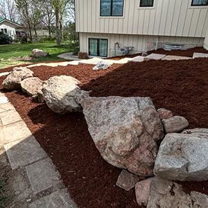 Landscaped yard with large rocks, red mulch, and a stone walkway beside a house