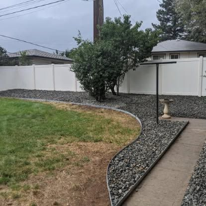 Curved gravel garden bed beside a concrete path and white fence, with a shrub and lawn under cloudy sky