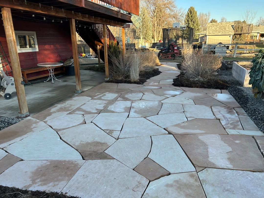 Stone patio beside a red house, with a cracked flagstone path and bushes in a yard.