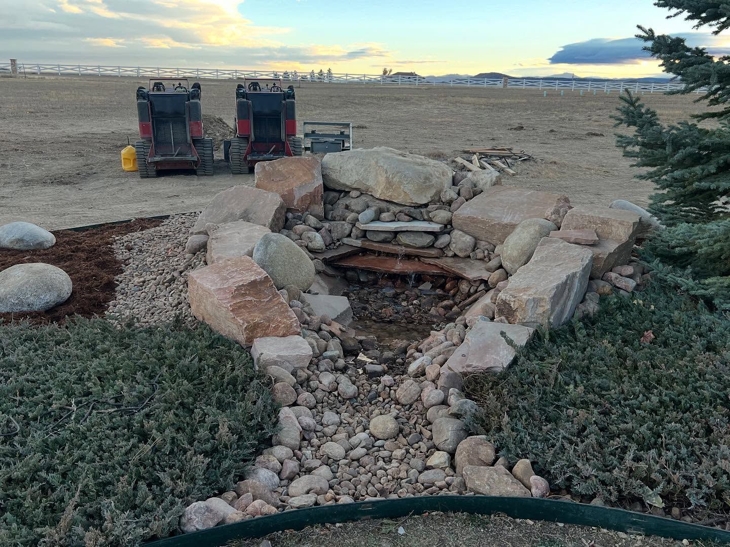 Stone fire pit in a grassy yard at sunset, with two grills in the background and mountains beyond.