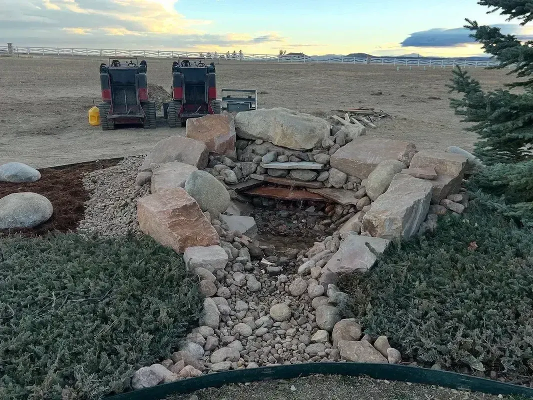 Stone fire pit in a grassy yard at sunset, with two grills in the background and mountains beyond.