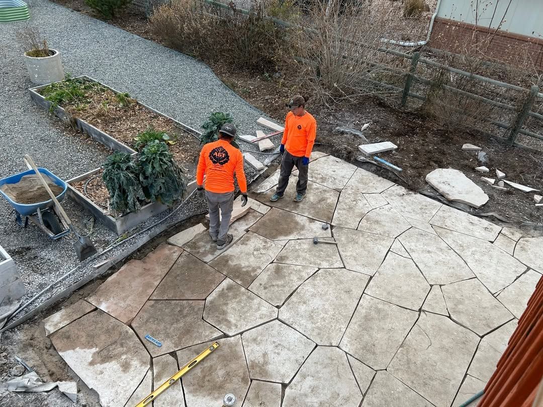 Two workers in orange jackets stand on a stone patio beside a gravel garden path.