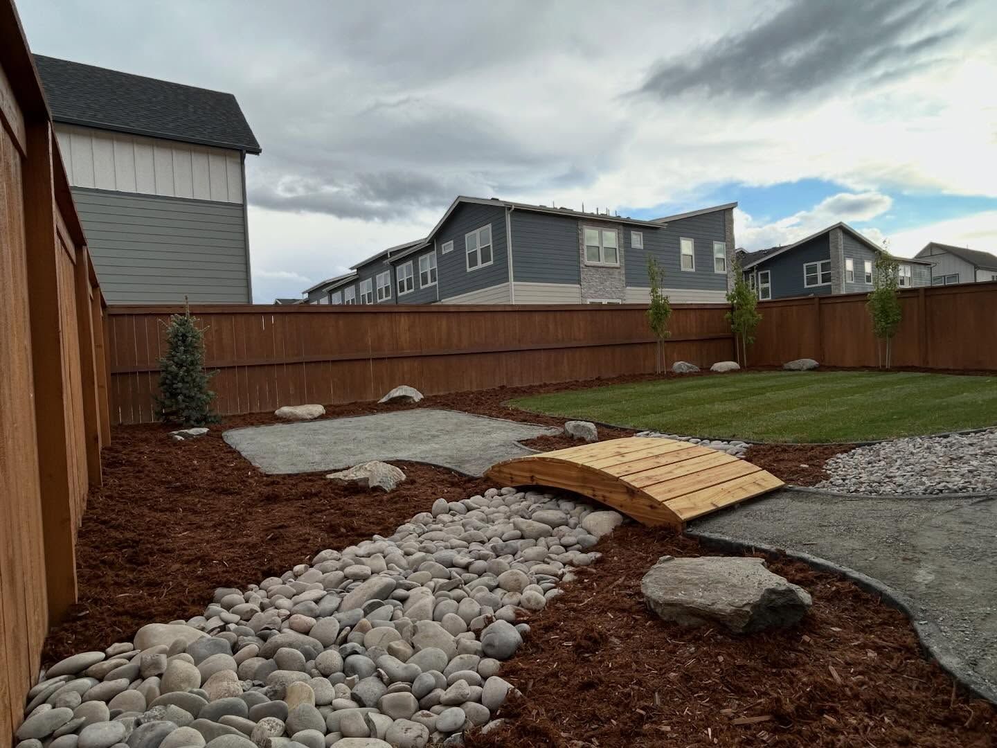 Backyard with gravel path, wood bridge over dry creek bed, and brown fence beside suburban homes