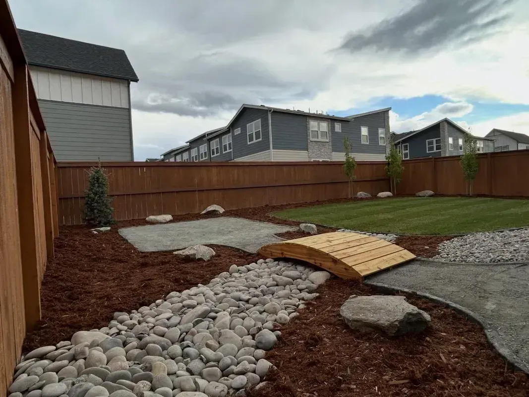 Backyard with gravel path, wood bridge over dry creek bed, and brown fence beside suburban homes