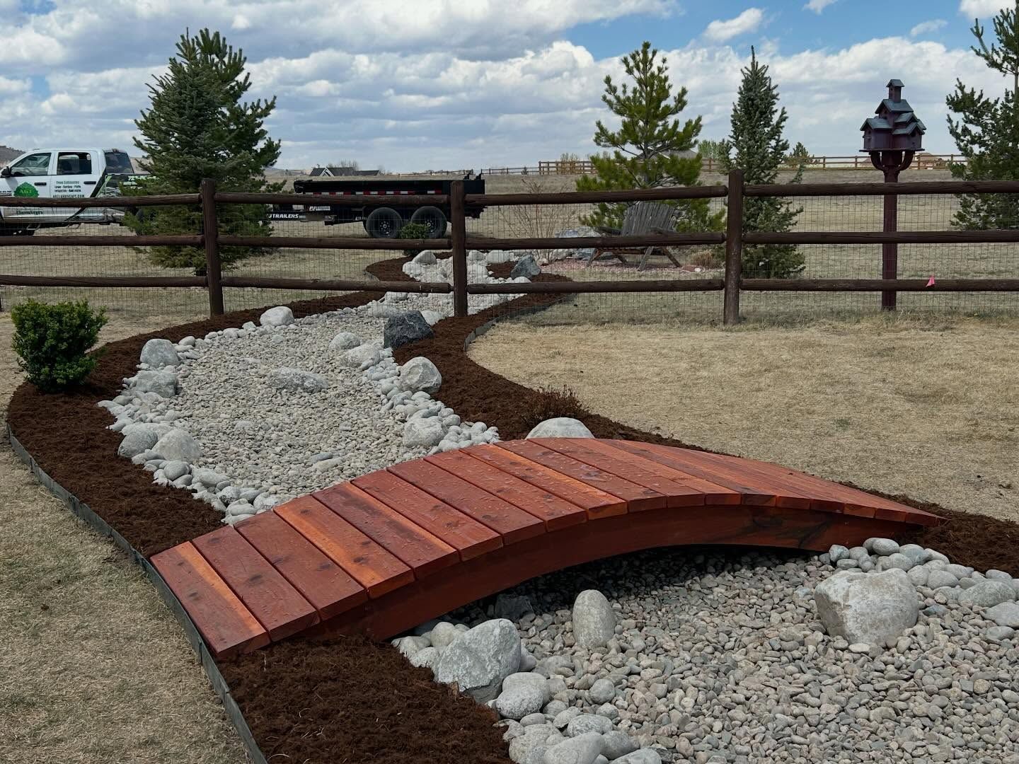 Curved red wooden footbridge over a dry rock-lined garden bed by a split-rail fence