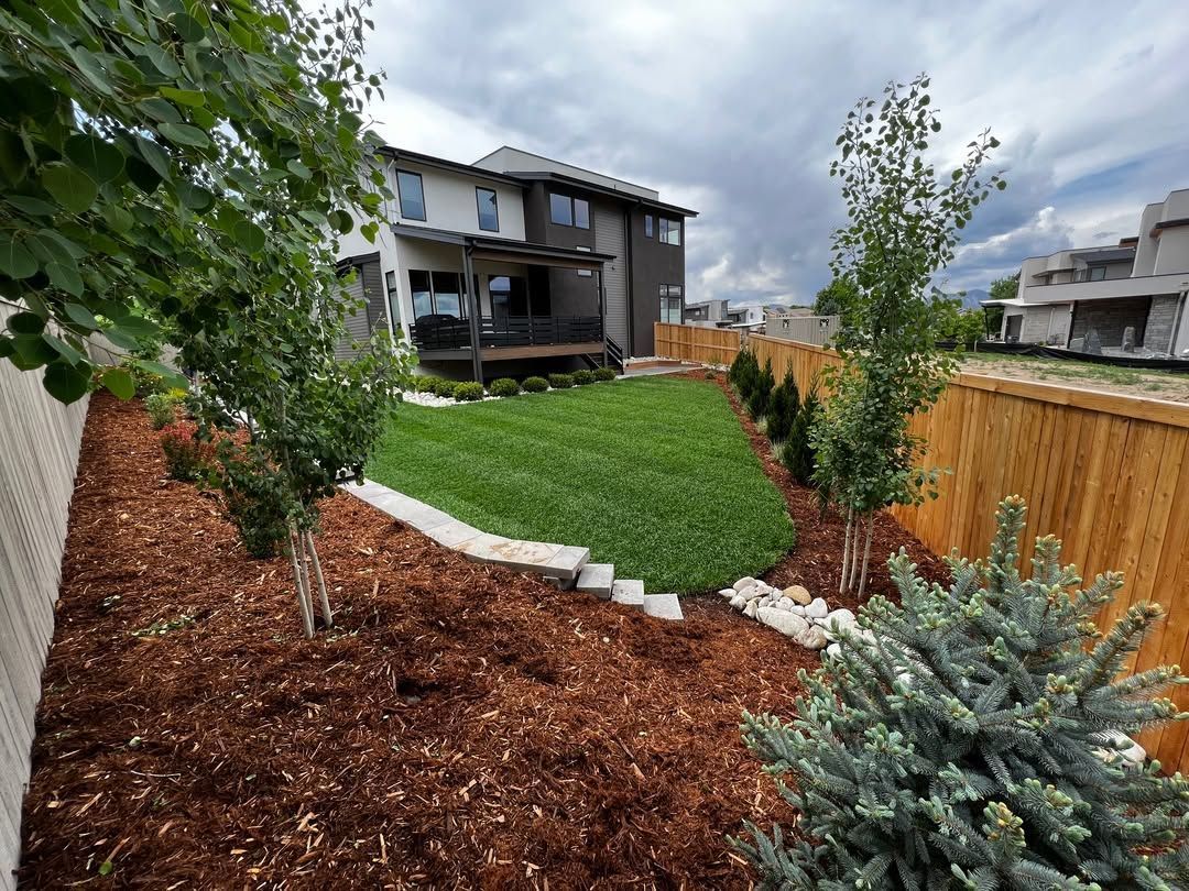 Landscaped backyard with green lawn, mulch beds, shrubs, and a two-story house in the background