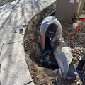 Worker crouching in a trench near a sidewalk, handling black pipes by a utility area.