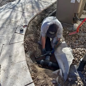 Worker crouching in a trench near a sidewalk, handling black pipes by a utility area.