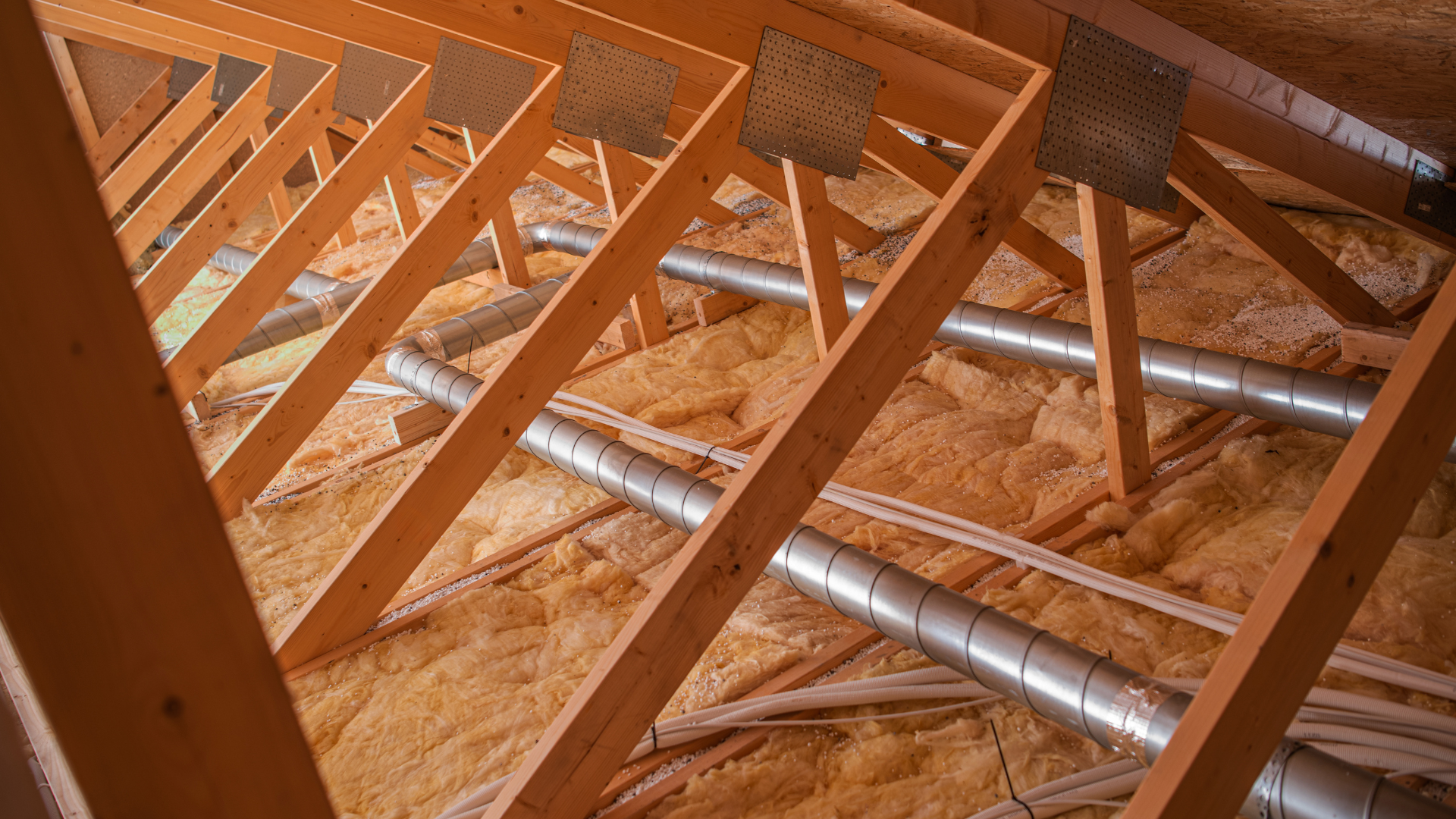 Technician in attic insulation suit vacuuming pink insulation between wooden rafters
