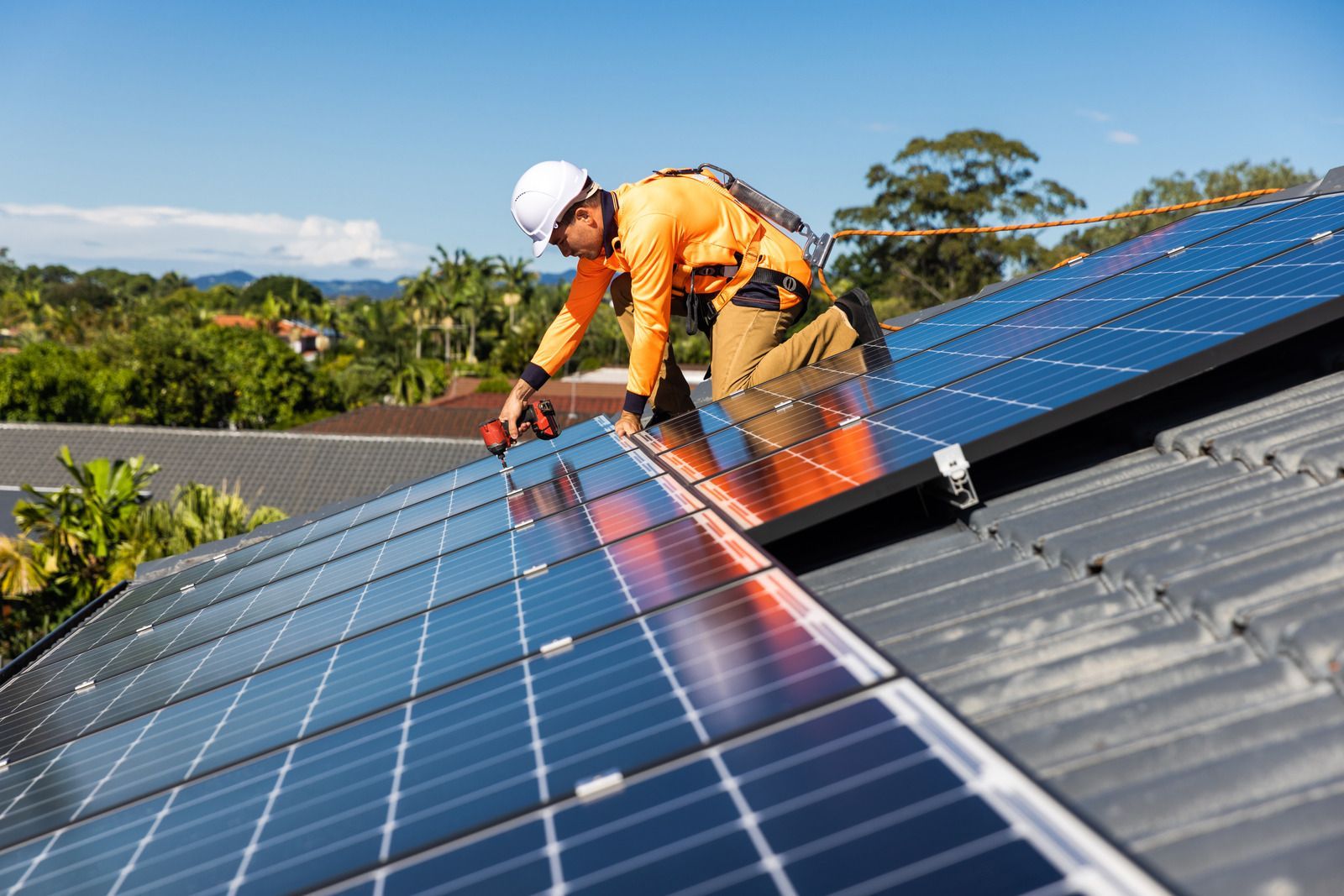 Roofer in orange jumpsuit installs solar panels on a rooftop on a sunny day.