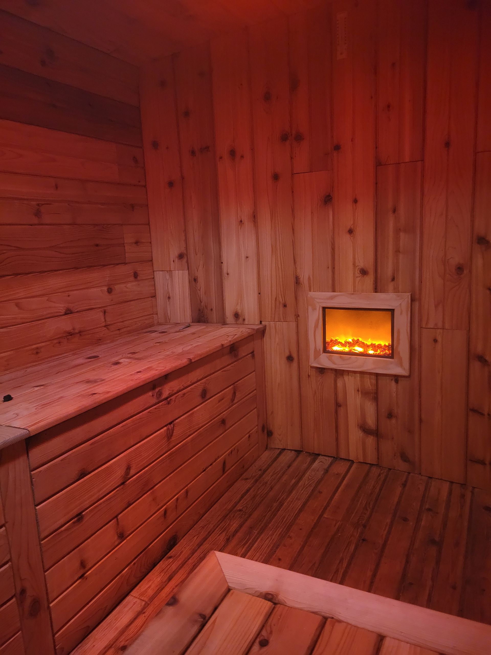 Wooden sauna interior, lit by red light. Bench and floorboards visible, fireplace feature on wall.