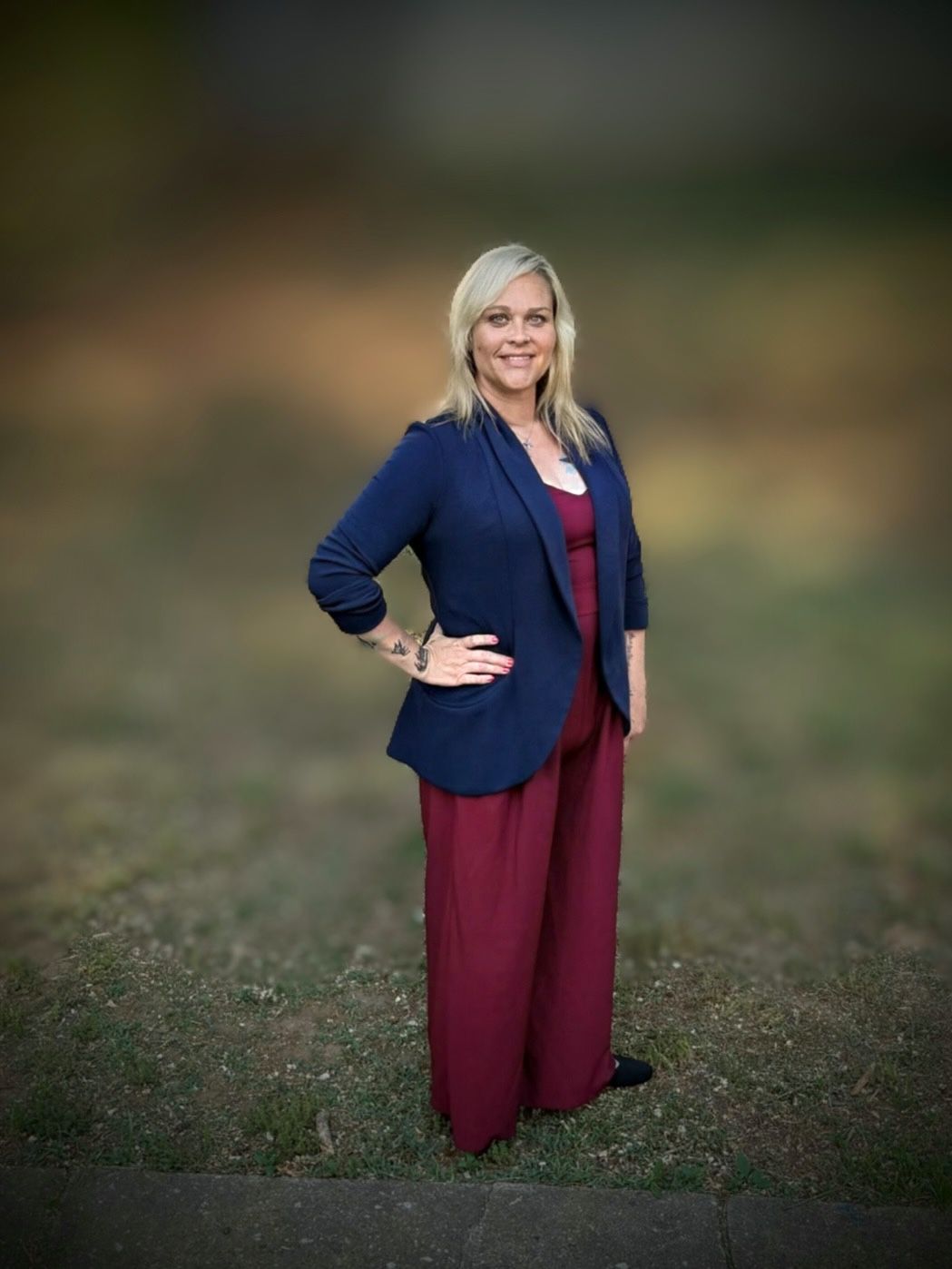 Woman standing outdoors on grass, wearing a navy blazer and burgundy outfit, with a blurred background.