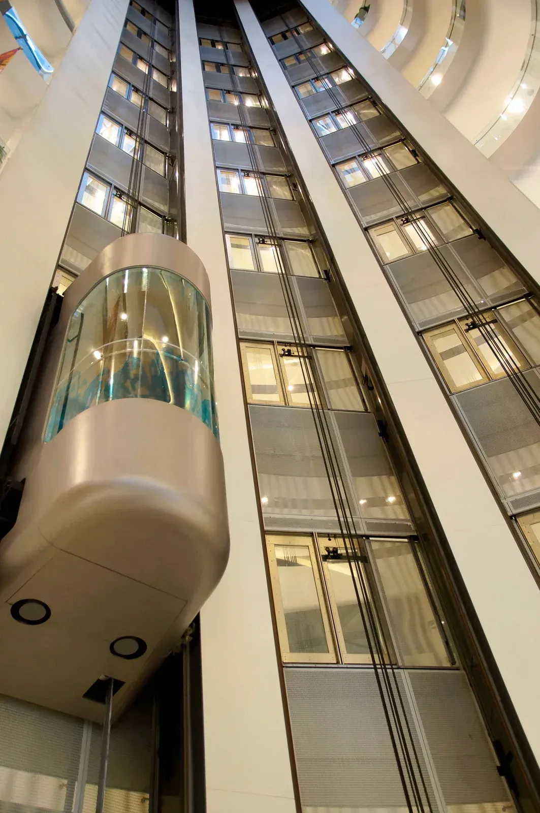 Elevator ascending in a tall building. Glass enclosure reveals interior. White walls, windows, and structural details visible.