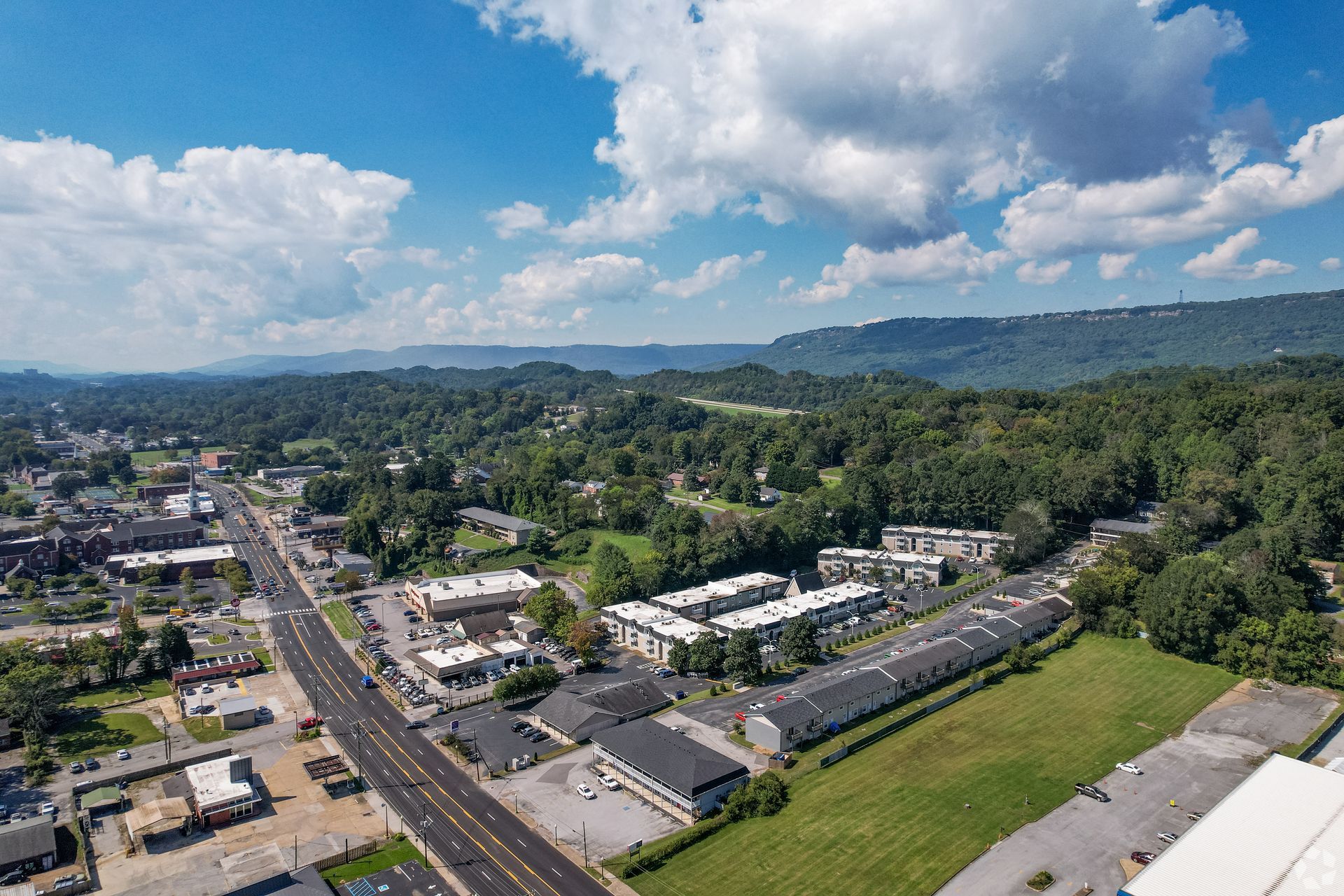 Aerial view of a small town with a main road, commercial buildings, apartment complexes, and mountains in the distance.