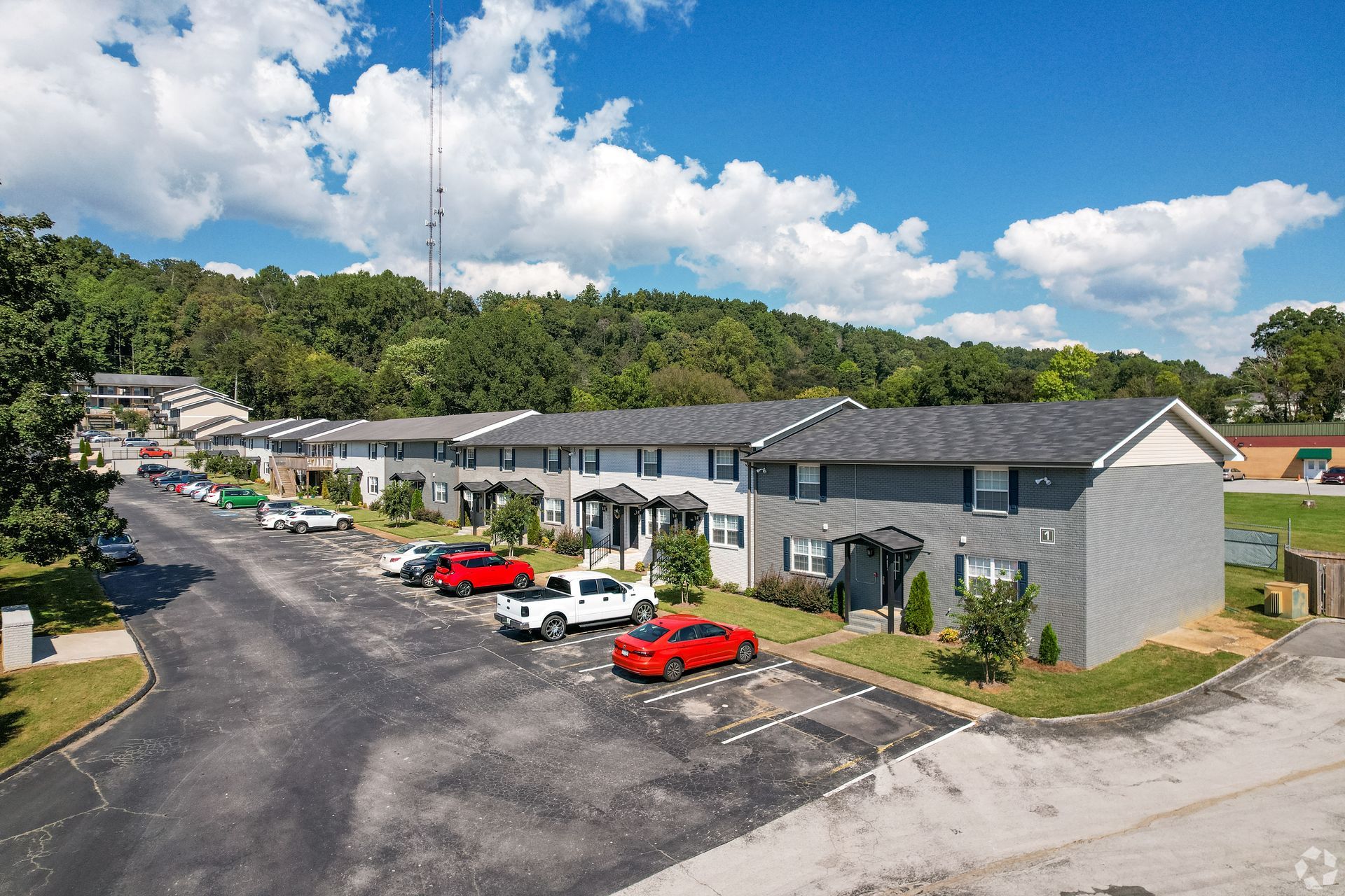 A row of gray apartment buildings with dark roofs, fronted by a parking lot with several cars, set against a wooded hill.