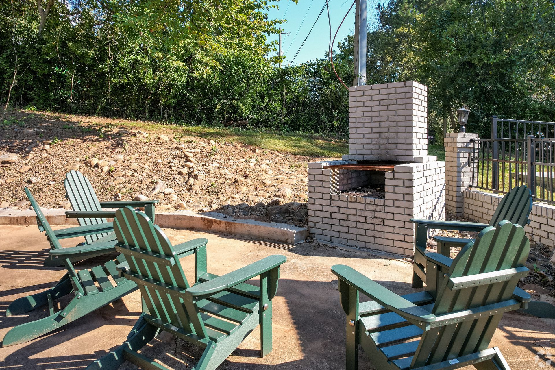 Four dark green Adirondack chairs arranged around a light-colored brick outdoor fireplace on a stone patio.