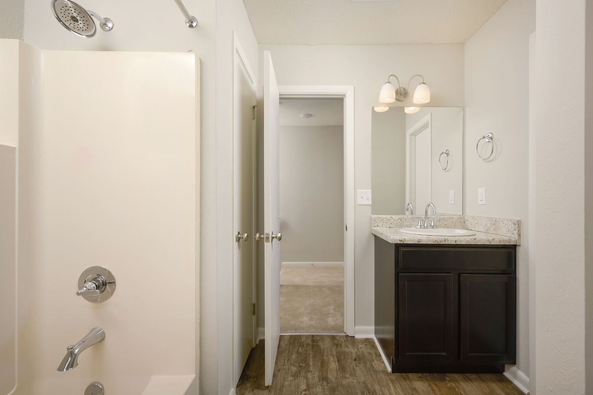 A bathroom with a white shower, a vanity with a dark cabinet and stone countertop, and an open door to a bedroom.
