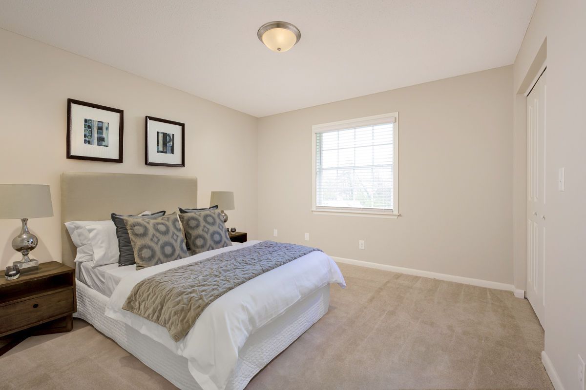 A neutral-toned bedroom features a bed with patterned pillows, a grey runner, two framed prints, and a light-filled window.