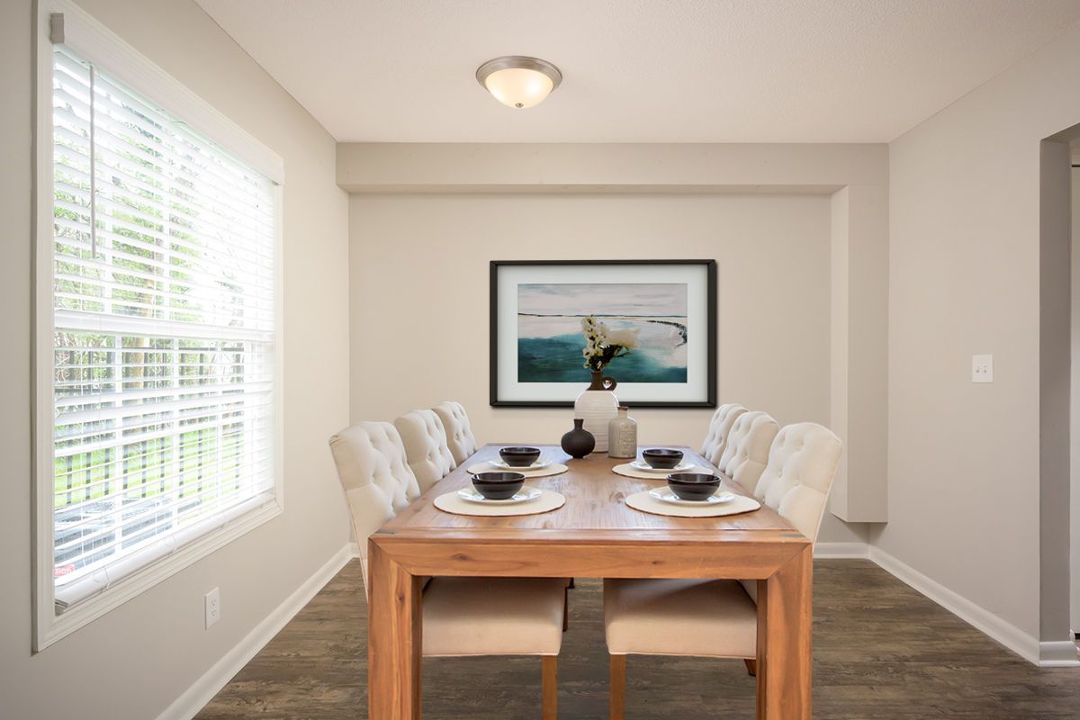 A dining room with a wooden table, six cream upholstered chairs, a landscape wall print, and a window with white blinds.