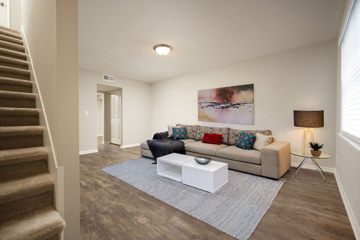 A living room with a beige sofa, white coffee table, patterned rug, abstract wall art, and stairs on the left.