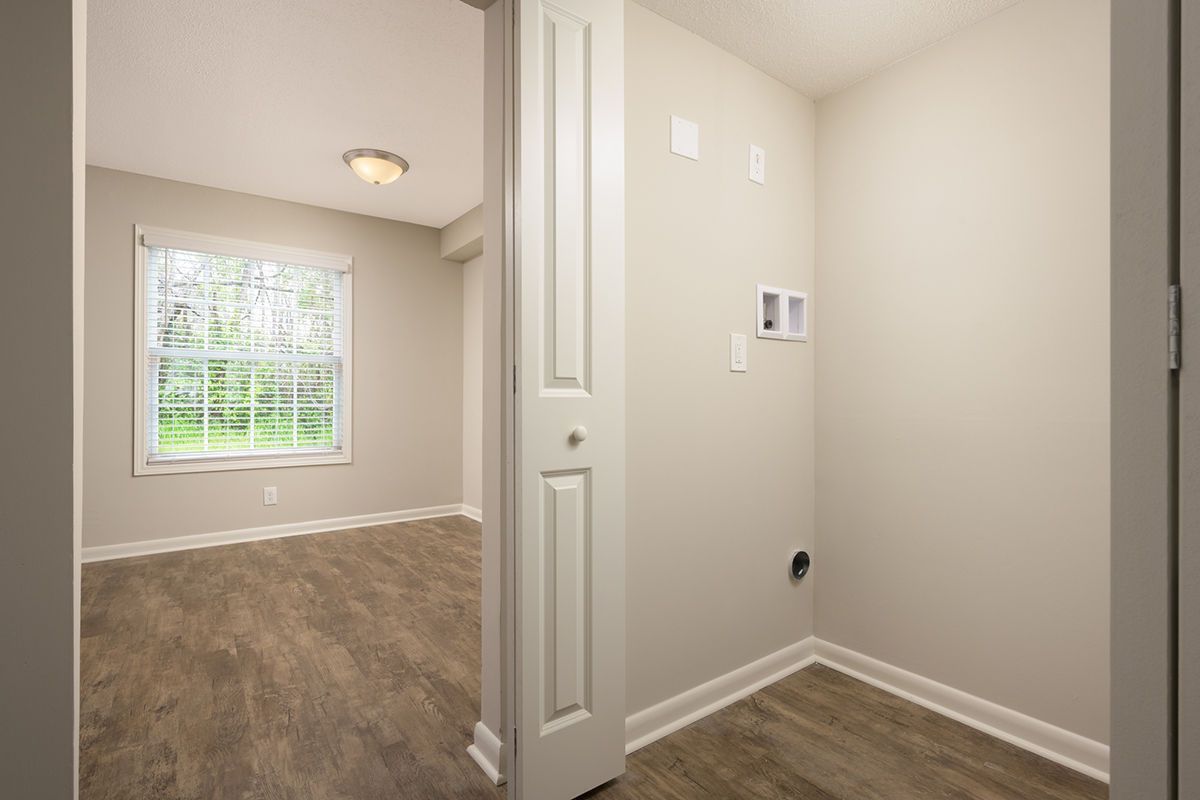 A view through a door into a room with a window, alongside an alcove with laundry hookups, all with light-colored walls.