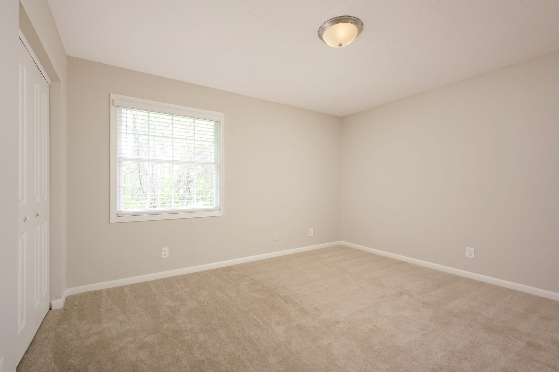 An empty room with beige walls and carpeting, featuring a window and a closed closet door.