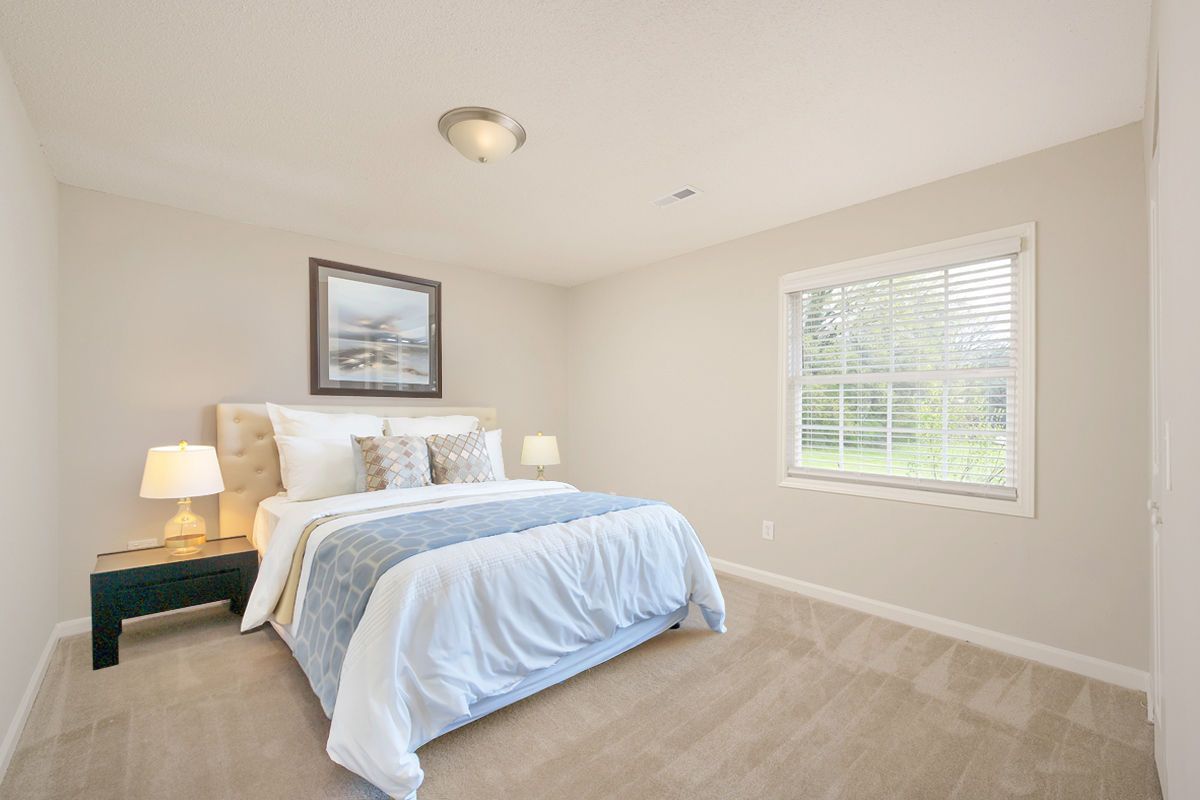 A bedroom featuring a bed with a blue throw, a framed picture, a lamp on a nightstand, and a window with blinds.