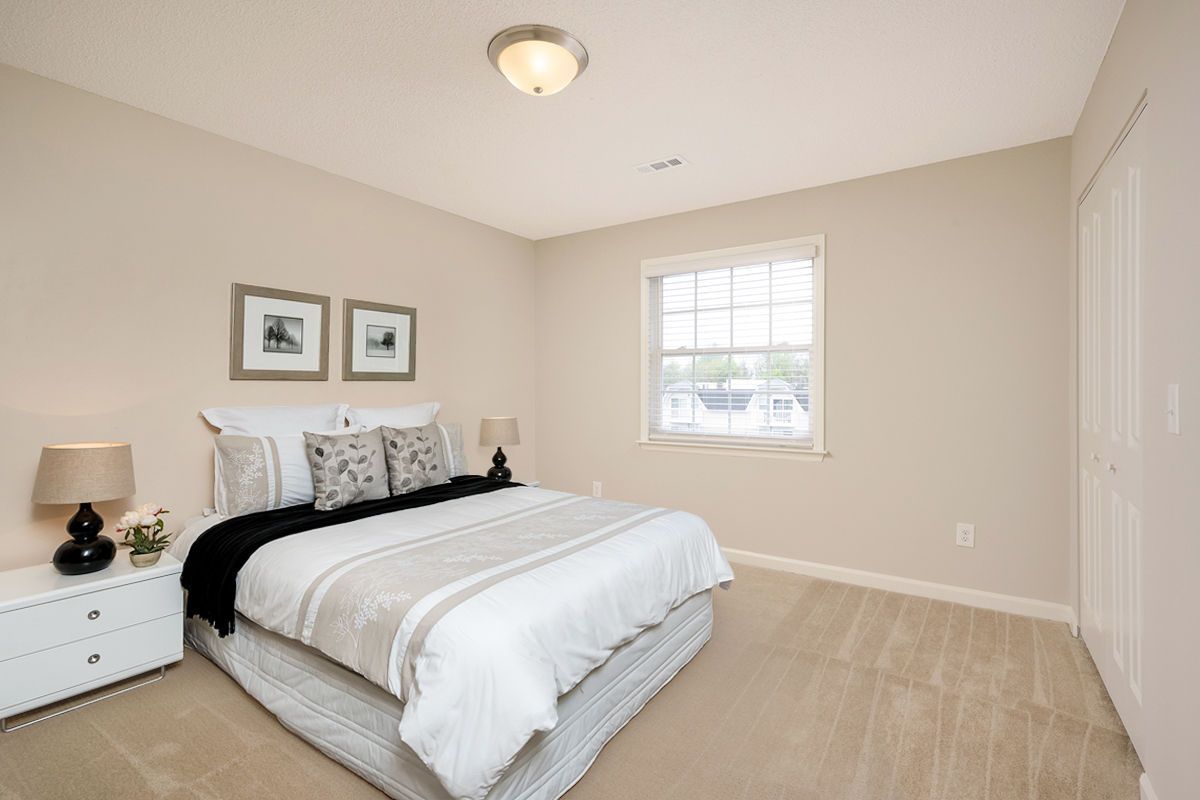 A neutral-colored bedroom featuring a queen bed with a white comforter, two framed pictures, and a matching nightstand.