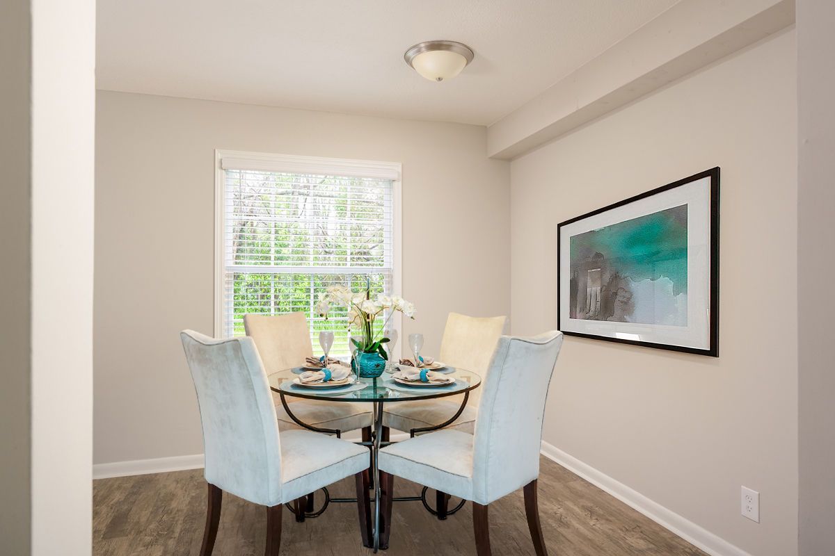 A round glass dining table with four light blue chairs sits in a room with light walls and a framed coastal print.