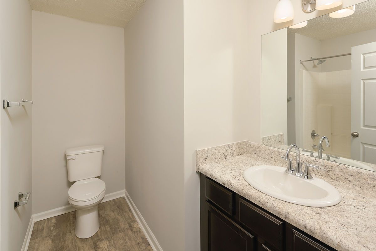 A bathroom featuring a light-colored countertop, dark wood vanity, white toilet, and a mirror reflecting a shower stall.