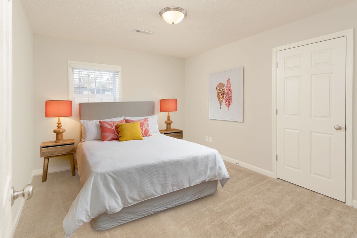 A bedroom with a queen bed, two orange bedside lamps, a window, beige carpet, and a white closet door on the right.