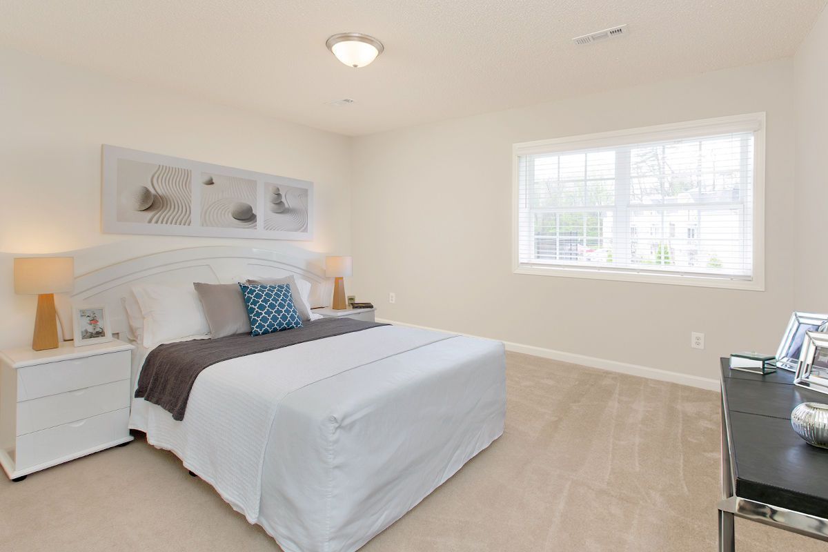 A bright, modern bedroom featuring a neatly made bed with white and gray linens, a white nightstand, and a window.