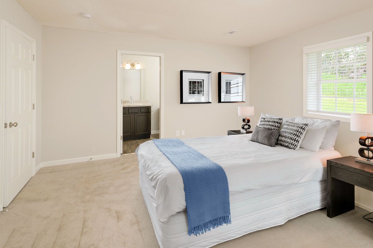 A bright bedroom with beige carpet, a bed with white linens and a blue throw, two wall framed photos, and an open doorway.