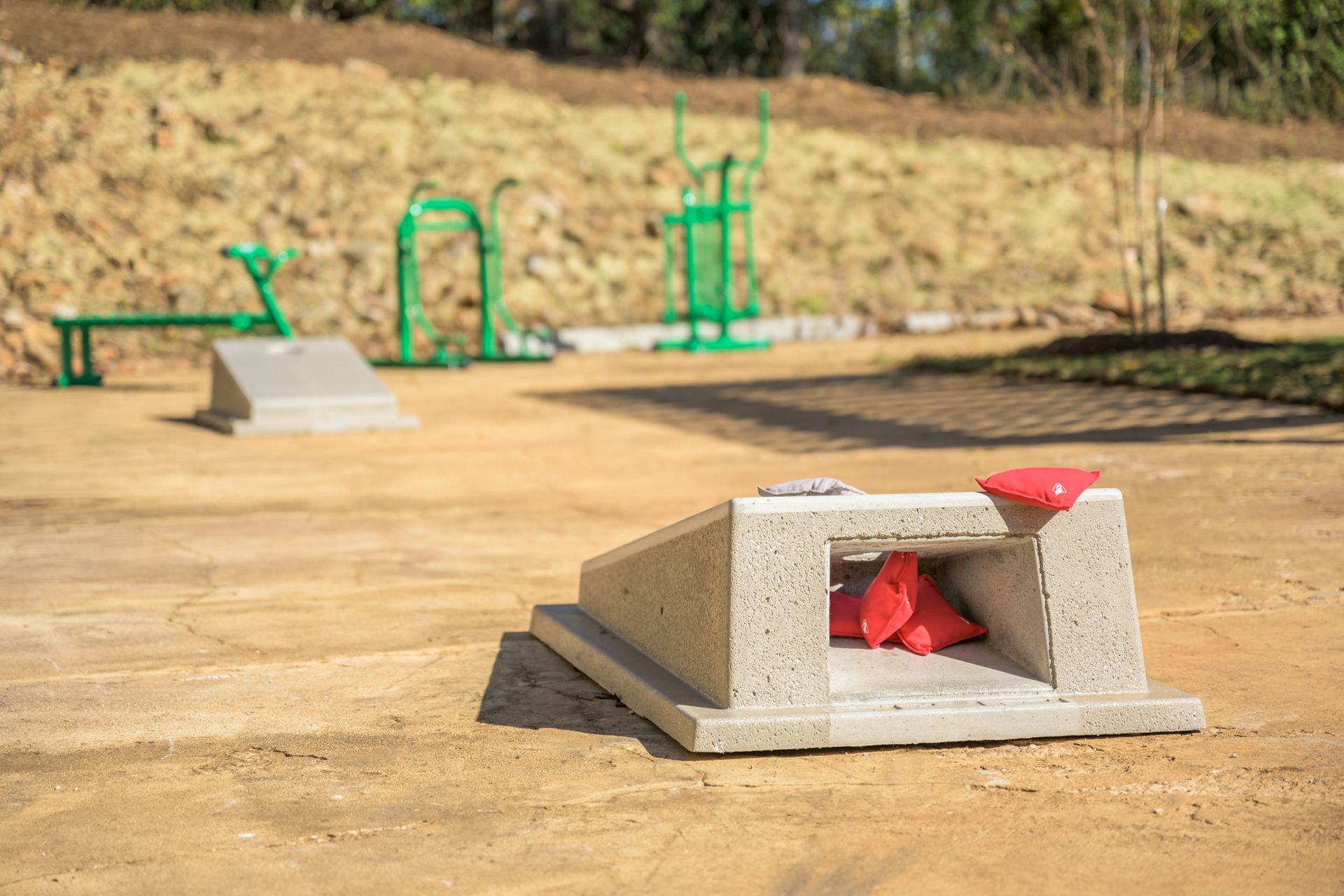 Cornhole boards with beanbags on a dirt ground in front of outdoor exercise equipment.