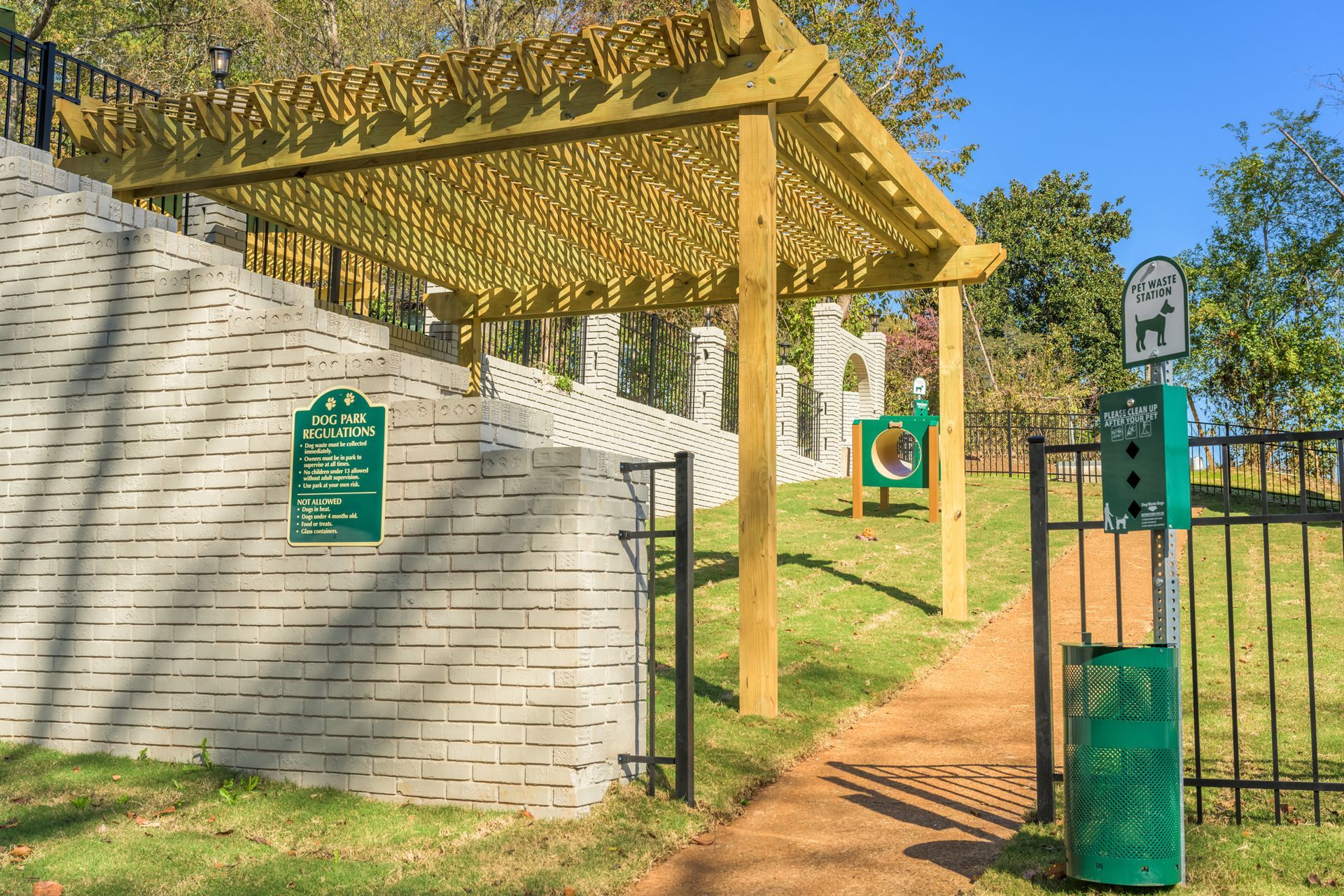 Entrance to a dog park with a wooden pergola, brick wall, signage, and a waste bag dispenser on a sunny day.