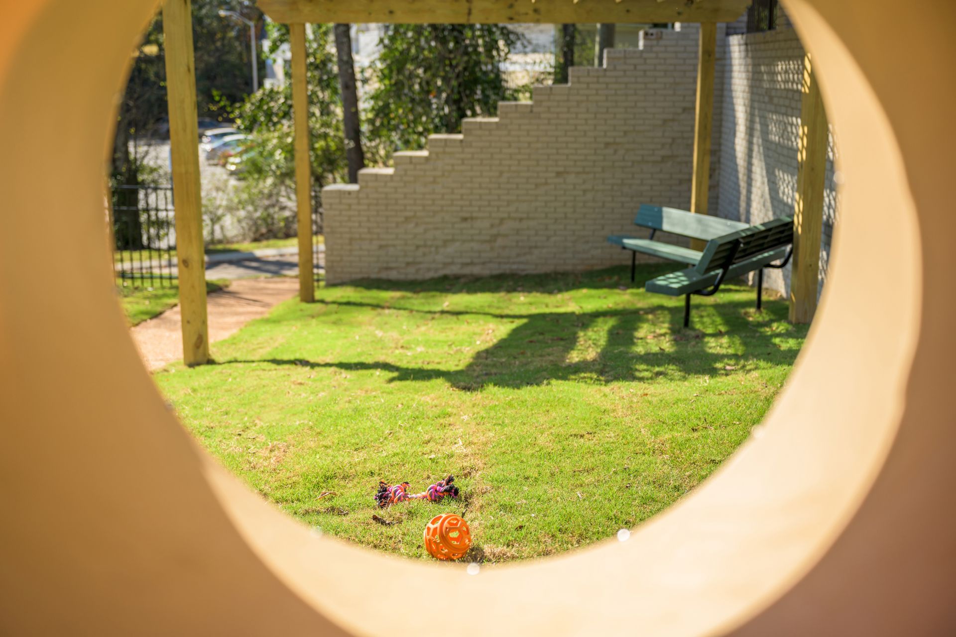 View through a circular opening showing a grassy park area with a pergola, a bench, and a discarded toy on the ground.