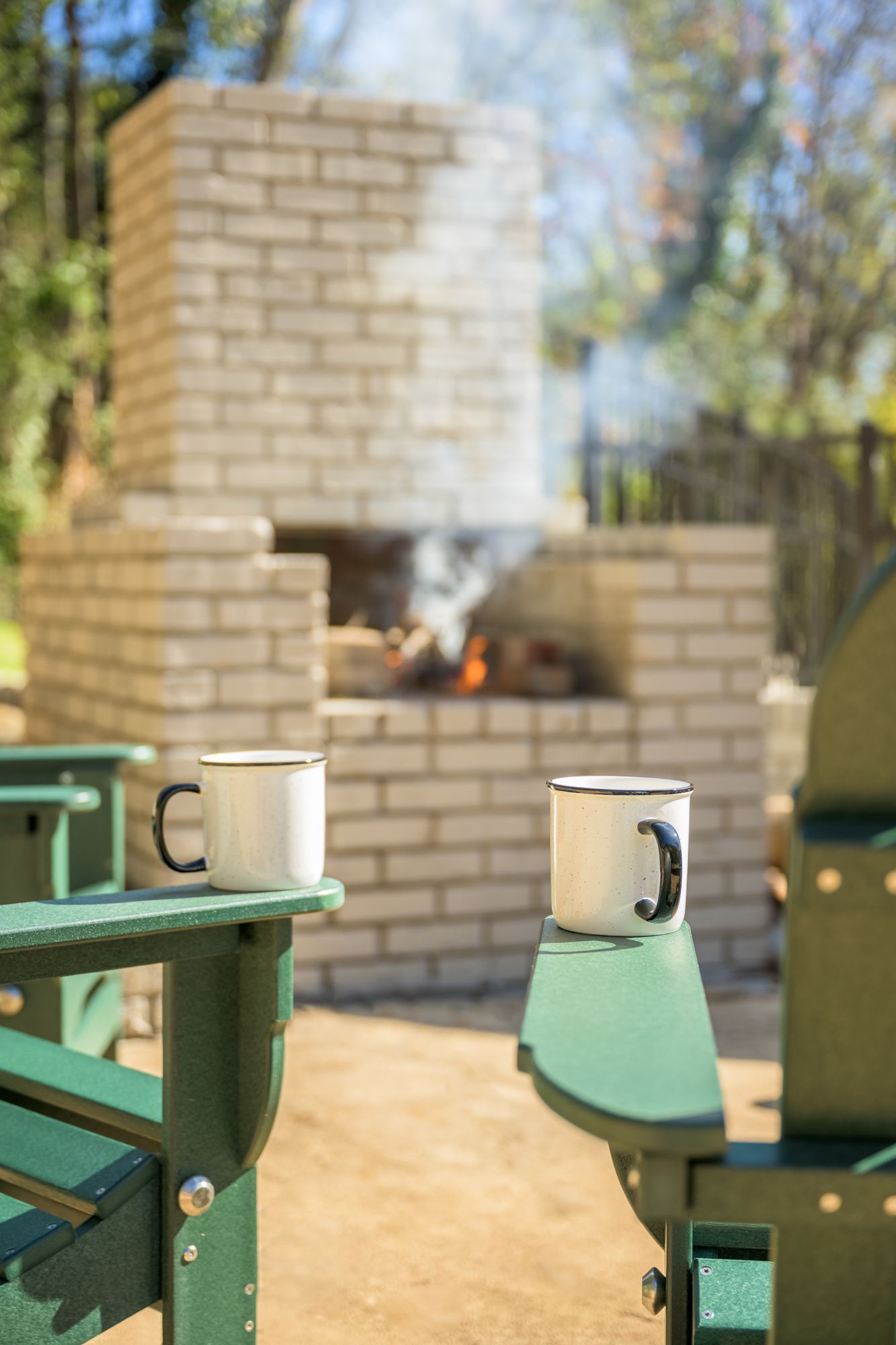 Two white mugs rest on the arms of green Adirondack chairs in front of a smoking brick outdoor fireplace.