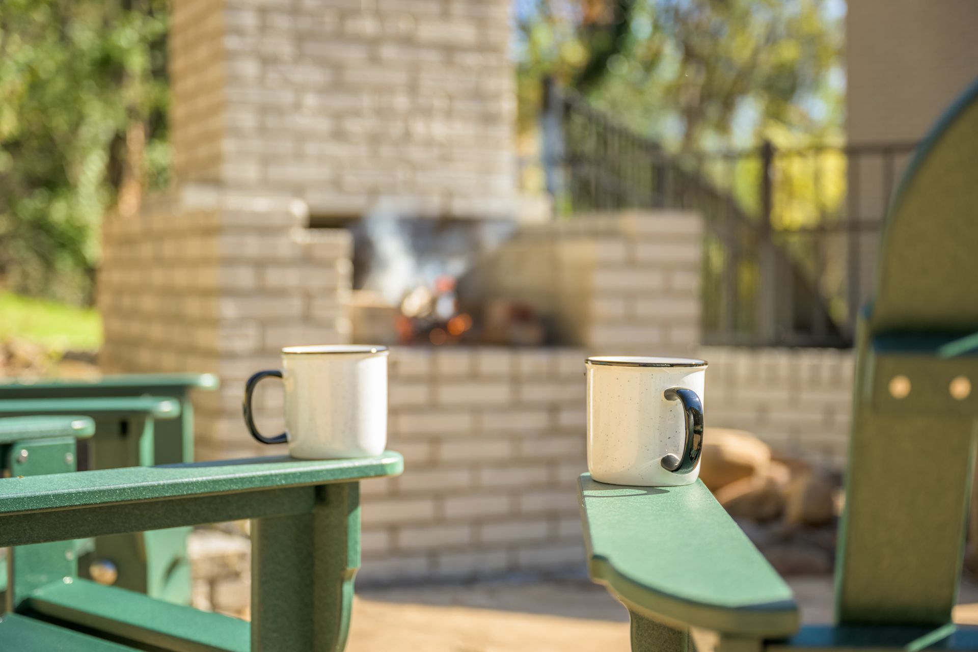 Two white mugs sit on the arms of green outdoor chairs in front of a stone fire pit.