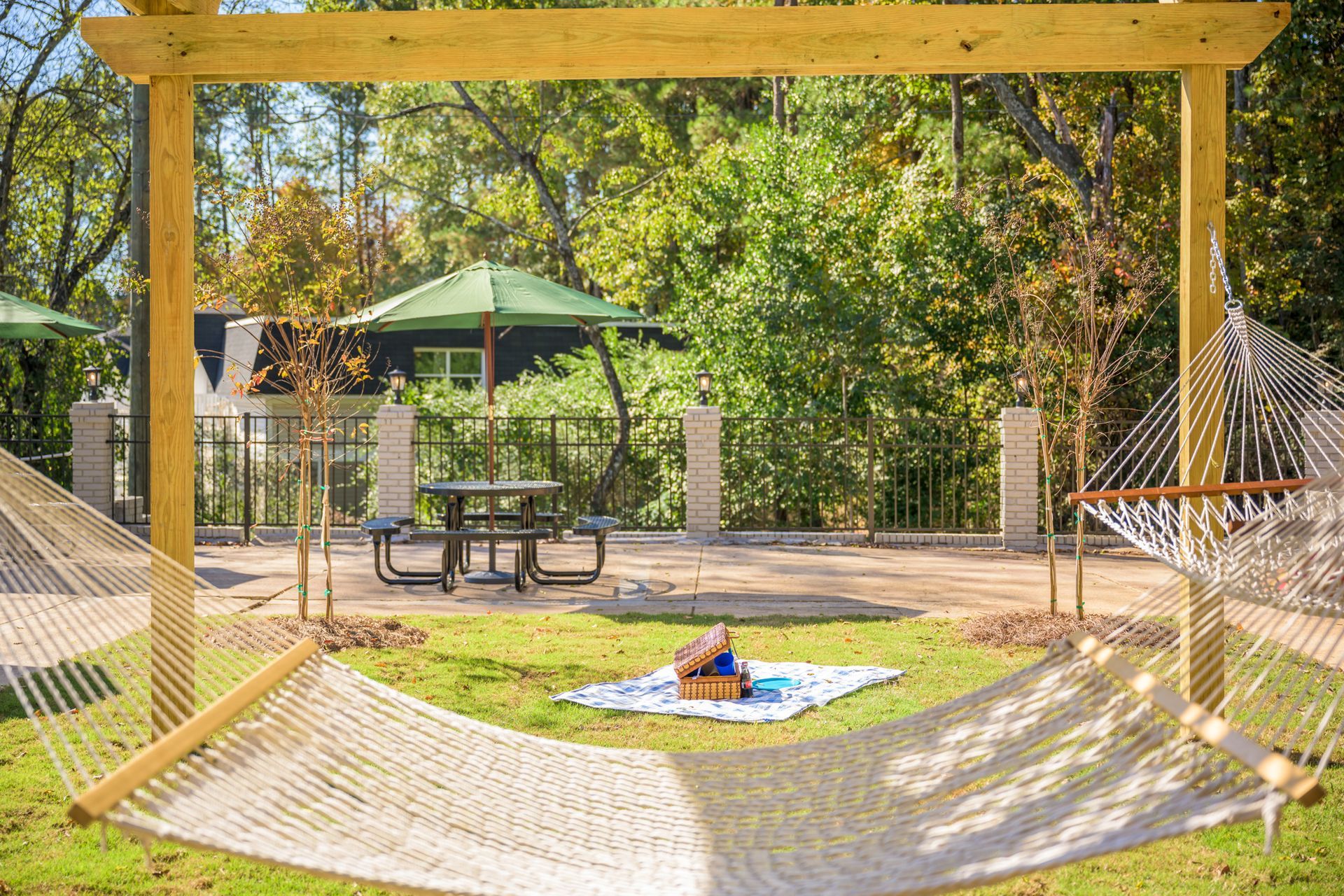 A rope hammock hangs in the foreground, framing a park scene with a picnic mat on the grass and a table under an umbrella.