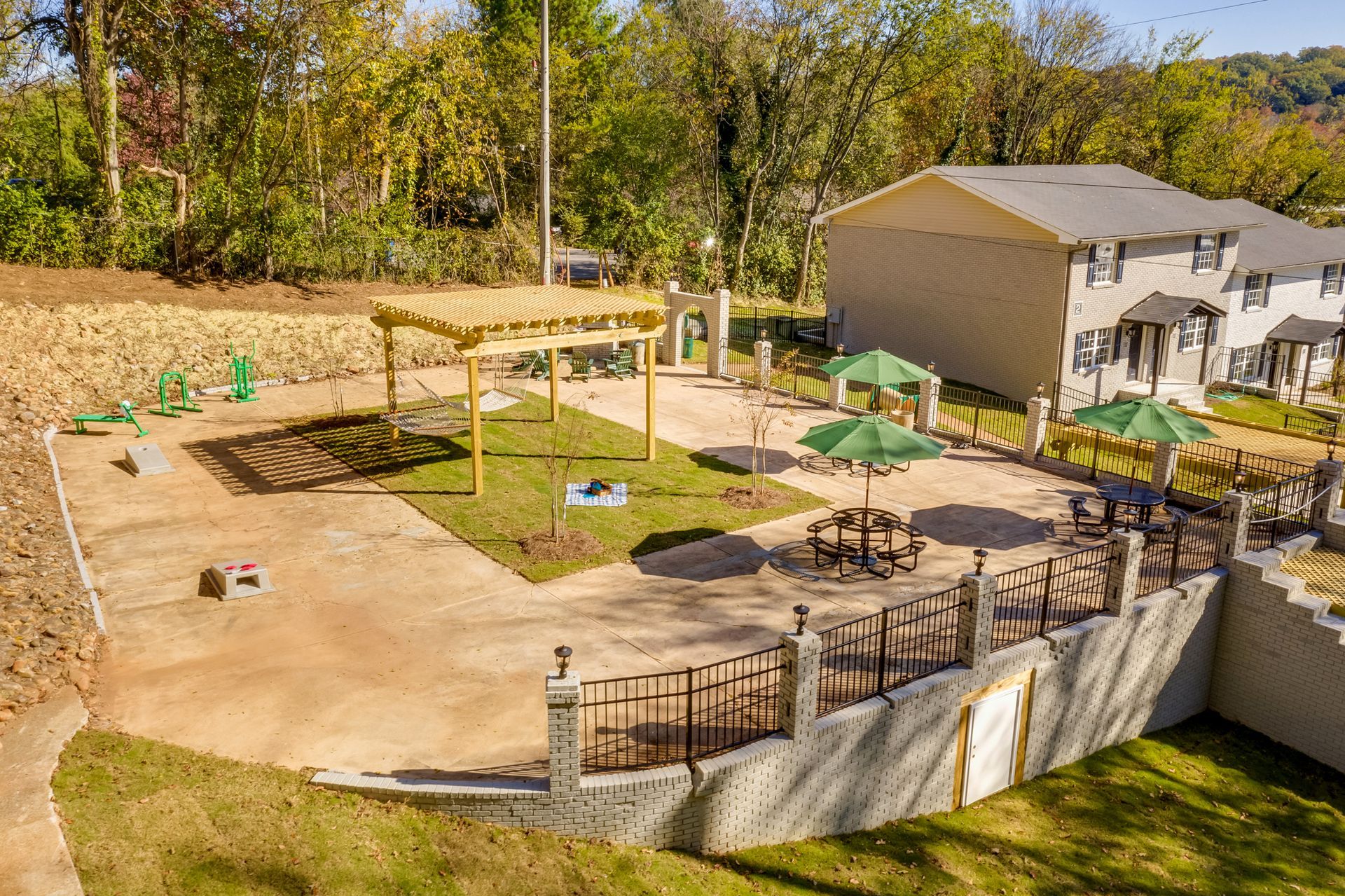 An outdoor patio with a wooden pergola, green umbrellas, picnic tables, and exercise equipment beside a two-story home.