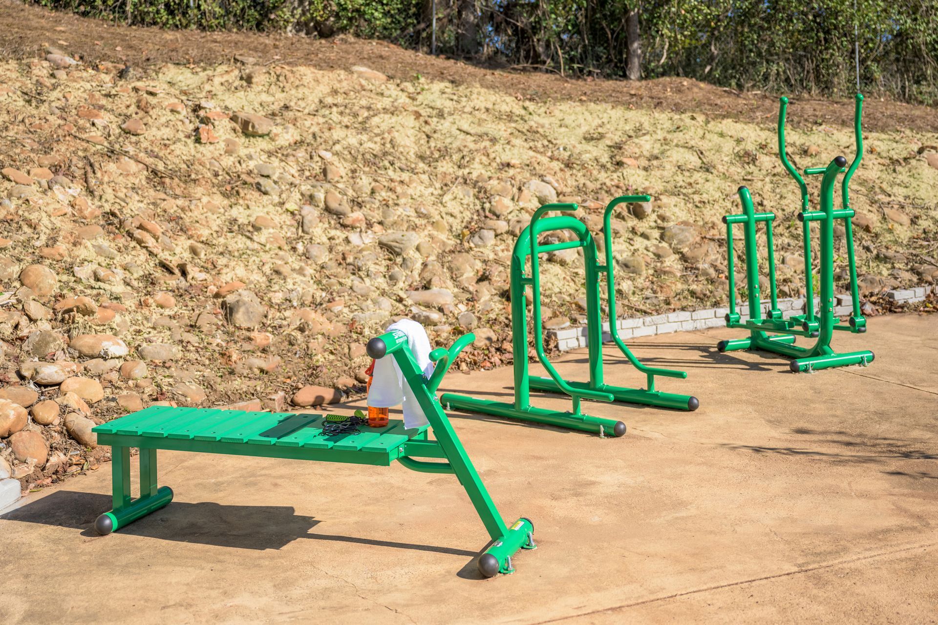 Outdoor fitness equipment, including a green bench and exercise machines, positioned on a concrete area against a hillside.